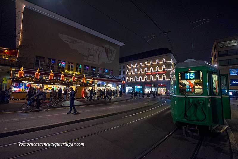 Mercadillo de navidad en Basilea, Imagen, un foto nocturna con un tranvía verde y sus railes. A la derecha unos puestos navideños con decoración de velas grandes con luces sobre el.