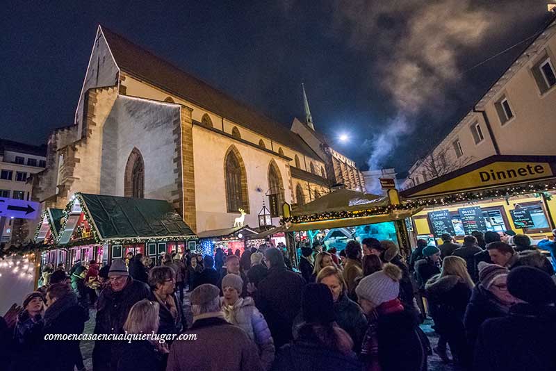 Imagen, en la calle una iglesia muy grande debajo puestos navideños y gente pasando. Al fondo hay un humo que parece que sale de una chimenea, trasmite frio.