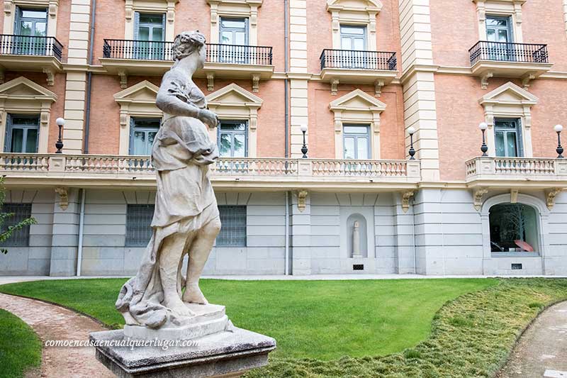 Imagen, en los jardines, una escultura a la izquierda de la foto, sobre un pedestal, de fondo las ventanas del edificio del museo.