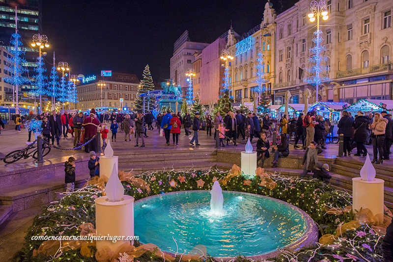 plaza Ban Jelačić o Josipa Jelacica. Imagen, se ve una plaza, abajo se ve una fuente azul con decoración navideña, esta decoración se ve tambien en las farolas en forma de pinos navideños. Se ve mucha gente. 