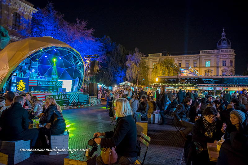 zagreb navidad. Imagen nocturna con un escenario en forma circular donde esta pinchando un dj. Hay gente sentada en cubos que hacen de sillas tomando algo y oyendo la música, al fondo un edificio institucional.