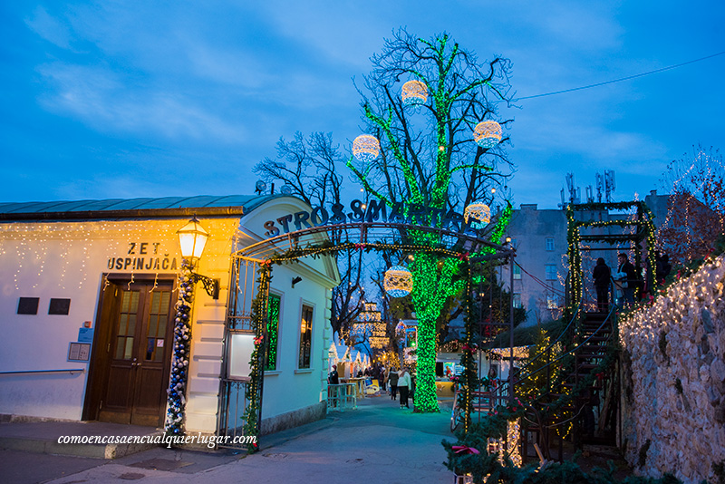 Plaza Strossmayer Zagreb. Imagen, es la entrada e tro mercadillo, en la hora azul, el cielo esta azul y en el centro se ve un arbol iluminado de verde con grandes bolas de navidad.