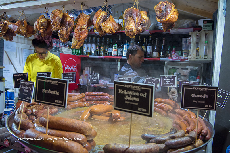 Croacia en Navidad. Un pequeño quiosco navideño en el que venden comida típica. En primer plano se ve un "sartén" muy grande con diferentes tipos de salchichas, dos personas trabajando, trozos de carne codillos, colgados y botellas de fondo. 