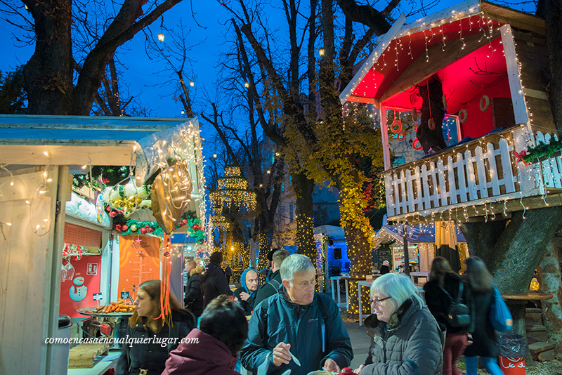 Croacia en Navidad, Foto en un plazo cerrado se ve unas casitas de madera con decoración navideña, a la derecha la casita esta en lo alto de un arbol. En la parte central de la foto se ven unas personas comiendo.  