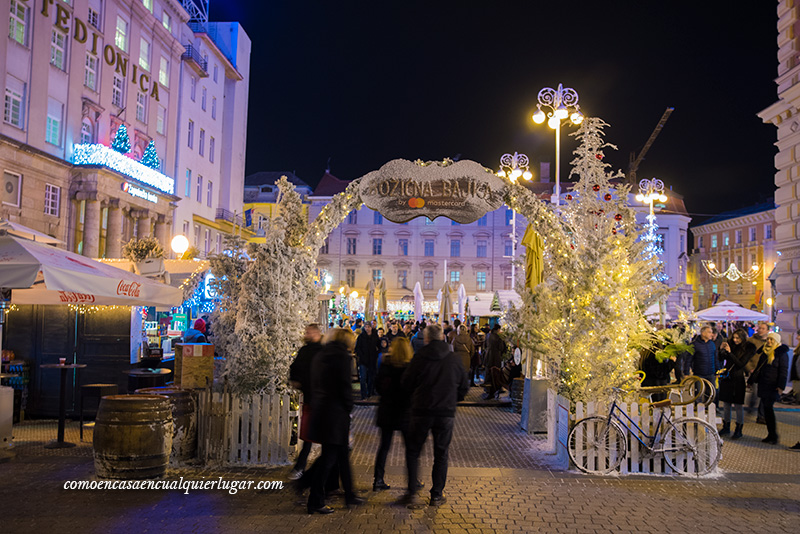 Croacia en Navidad. Imagen, se ve la entrada por un arco al mercadillo, el arco esta escrito el nombre del mercadillo y unos arboles de plástico en color blanco. Como decoracion hay un bicicleta en el lado derecho y gente entrando por debajo de los arcos.