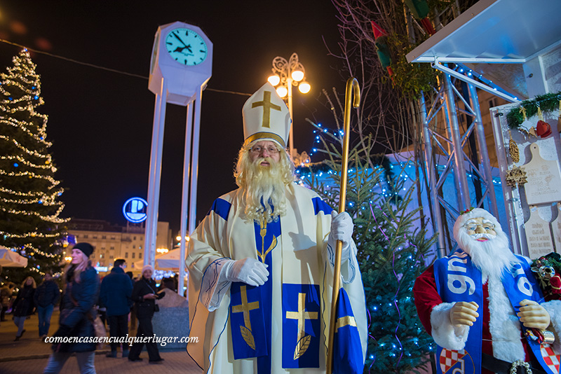 plaza Ban Jelačić o Josipa Jelacica. Imagen, un retrato de medio cuerpo de un señor vestido como papa noel, con largas barbas, lo curioso es que el traje que lleva es azul y blanco. lleva un gorro de obispo llamado solideo con una gran cruz amarilla simulando oro.En el fondo se ven una gran torre con un reloj.