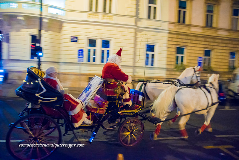 Imagen, un carro de paseo tirado por dos caballos blancos. El conductor es santa claus y de pasajero va la señora se santa.