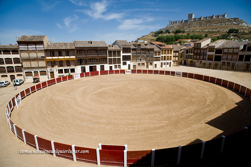 Imagen, plaza de coso, por la luz parece que el suelo es amarillo. Se ven las casa antiguas alrededor de la plaza y de fondo el castillo, en lo alto.