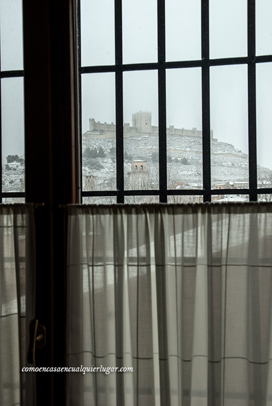 Imagen, desde un interior de una casa y mirando por una ventana con barrotes se aprecia un día de nieve, blanco, con el castillo de fondo.