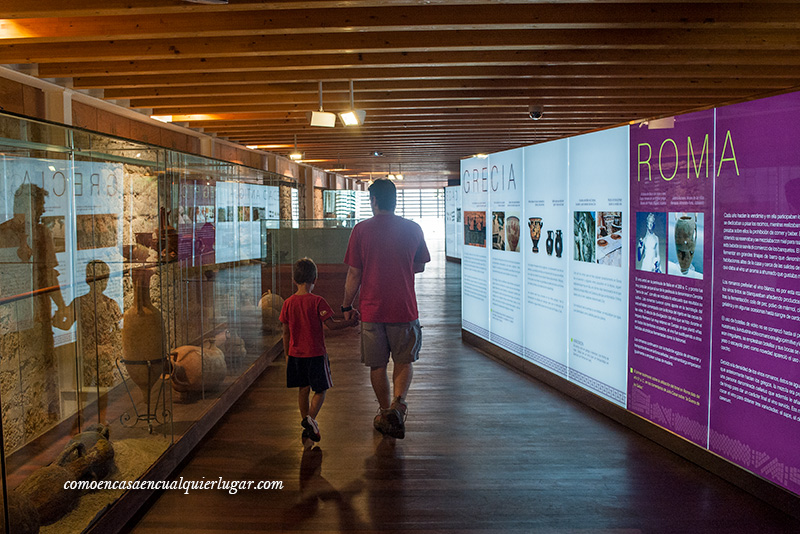 Imagen, un padre y un niño van de la mano andando por los pasillos del museo. En los pasillos se ve información de Grecia y Roma.
