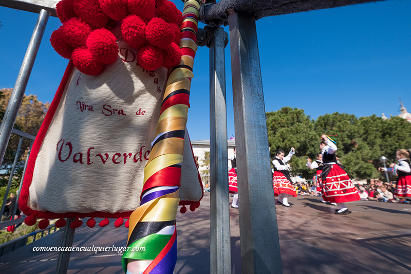 imagen, un zurron en el que esta bordado las letras de nuestra señora de valverde y de fondo unas niñas realizando bailes tradicionales.Santuario de Nuestra Señora de Valverde