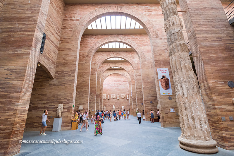 Foto de la sala principal, en la que se ven los arcos de piedra de su arquitectura, con una columna a la derecha muy grande  que llega hasta el techo y gente visitando la exposición 