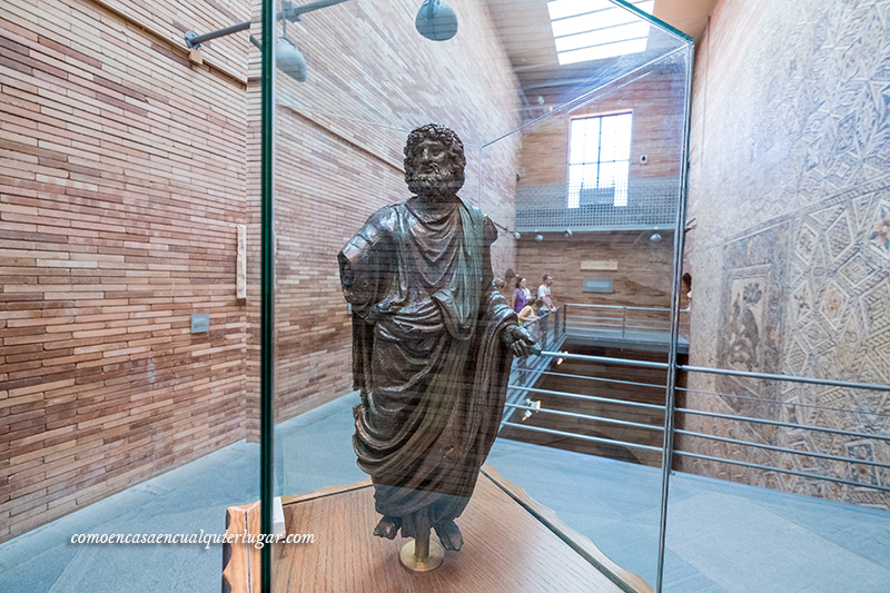 Foto, una estatua romana con una túnica, en la que se puede apreciar las pliegues de la tela en la piedra, sin brazo derecho. Esta metida en una urna de metacrilato que visitar en Mérida