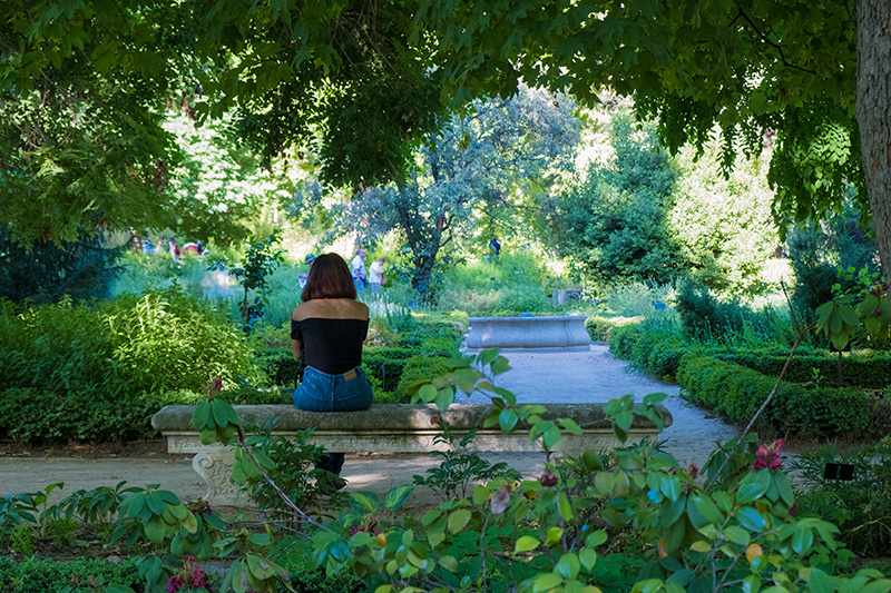 Foto. una mujer esta sentada en un banco de piedra, alrededor todo vegetación y una fuente de lejos